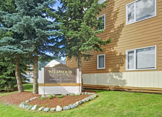 Sign for Wildwood Estates Apartment Homes, surrounded by lush green grass and tall pine trees, next to a tan building with multiple windows.