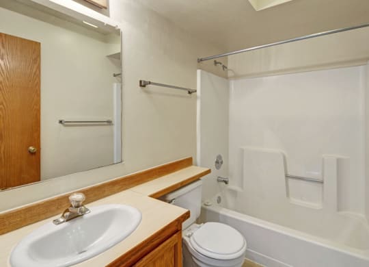 Small, tidy bathroom here at Wildwood Estates Apartments with white fixtures. A sink with wooden cabinet, toilet, and bathtub with shower inside are visible. Neutral color tones.