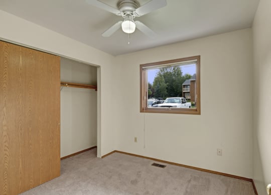 Small empty bedroom here at Wildwood Estates Apartments with beige carpet, white walls, and a wooden closet. A window shows parked cars and greenery outside. A ceiling fan is centered above. Cozy ambiance.