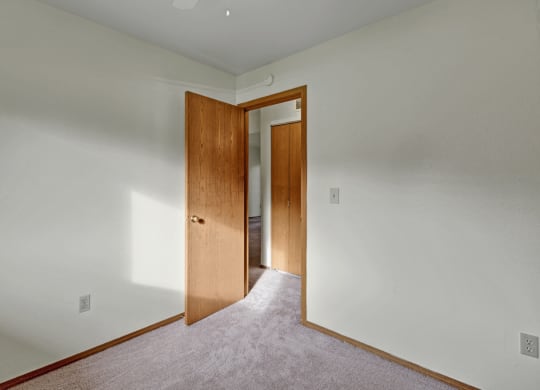 Empty bedroom here at Wildwood Estates Apartments with off-white walls, beige carpet, and an open wooden door. Sunlight casts soft shadows, creating a calm and inviting atmosphere.