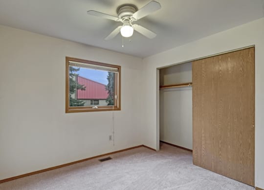 Plain bedroom here at Wildwood Estates Apartments with beige carpeting and white walls, featuring a ceiling fan. A window shows a view of a tree and red-roofed building. Open closet with wooden sliding door.