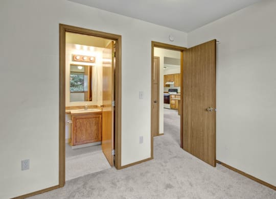 A beige carpeted room here at Wildwood Estates Apartments with two doors; one leads to a lit bathroom featuring a wooden vanity, and the other to a kitchen with wooden cabinets.