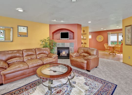 Warm living room here at Wildwood Estates Apartments with yellow and red walls, featuring leather sofas, a glass coffee table, fireplace, and a dining area with wooden chairs in the background.