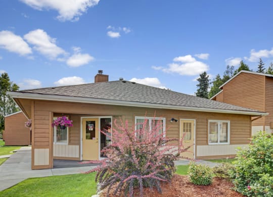 A single-story, brown wooden house here at Wildwood Estates Apartments with a grey roof sits under a bright blue sky. Pink flowers and green shrubs adorn the front, adding a welcoming touch.