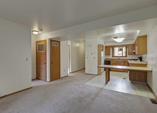 Spacious, empty living area here at Wildwood Estates Apartments with beige carpet, wood panel doors, and an adjoining kitchen. The kitchen features wooden cabinets and vinyl flooring, creating a simple, neutral tone.