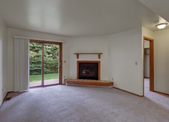 Living room here at Wildwood Estates Apartments with a cozy fireplace, white walls, grey carpet, and a sliding glass door opening to a lush green garden, creating a serene atmosphere.