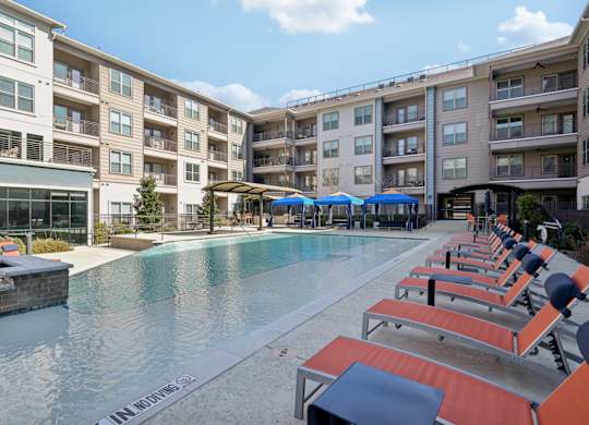 A pool surrounded by sun loungers and umbrellas in front of apartment buildings.