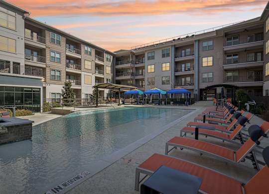 A swimming pool surrounded by sun loungers and umbrellas in front of apartment buildings.
