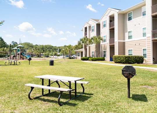 A picnic table is in the foreground of a grassy area with apartment buildings in the background.