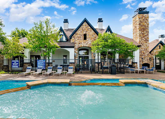 Pool with water feature in front of stone-accented clubhouse and poolside lounge chairs.