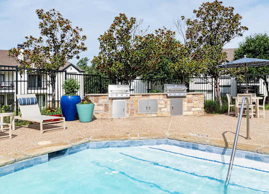 Poolside grilling area with two barbecue stations, lounge seating, and shaded outdoor table.
