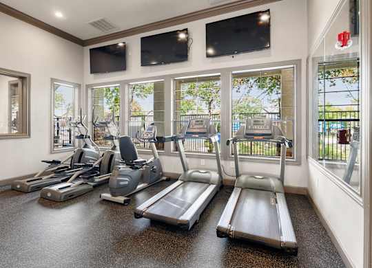 Fitness center with treadmills, bikes, ellipticals, tall windows, and wall-mounted televisions.