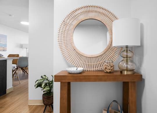 A round mirror with a wooden frame is on a table with a white lamp and a plant.