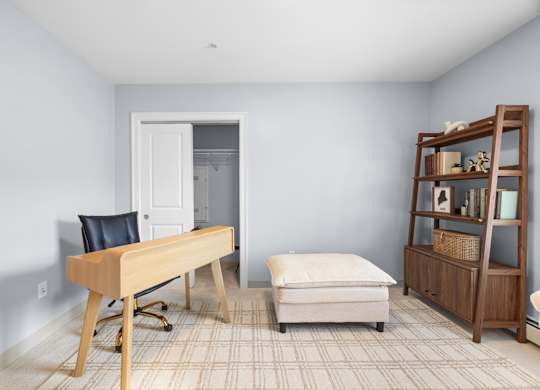 A room with a chair, a desk, a bookshelf and an ottoman