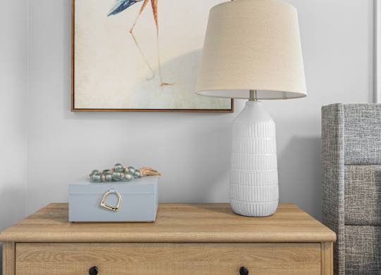 A table with a blue box, white lamp and artwork hanging on the wall behind it.