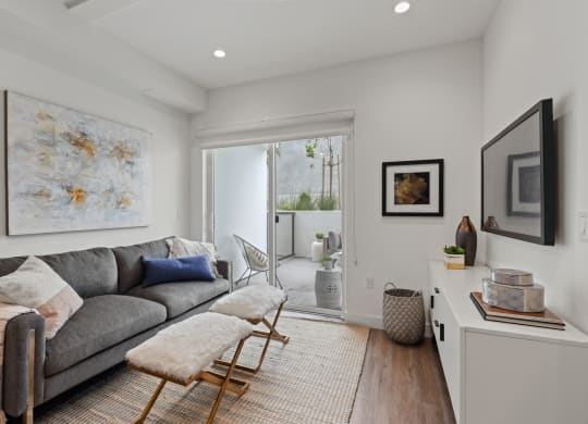a living room with a gray couch and two stools in front of a sliding glass door at The Fairfax Flats Apartments, Los Angeles, CA
