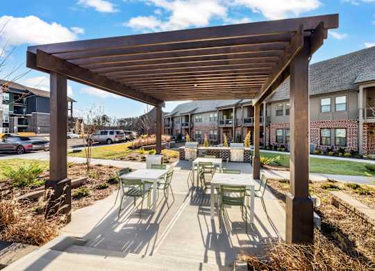 A wooden pergola with green chairs and tables is in the foreground of a residential area.