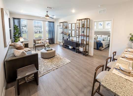 A well-lit, spacious living room with a large window, a sofa set, and a dining area with a table set for two.