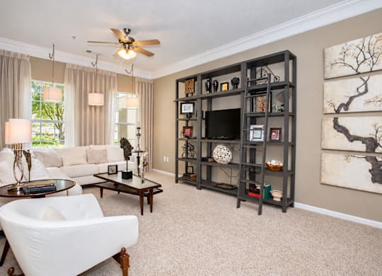 A living room with a white couch, a coffee table, a ceiling fan, and a large bookshelf.