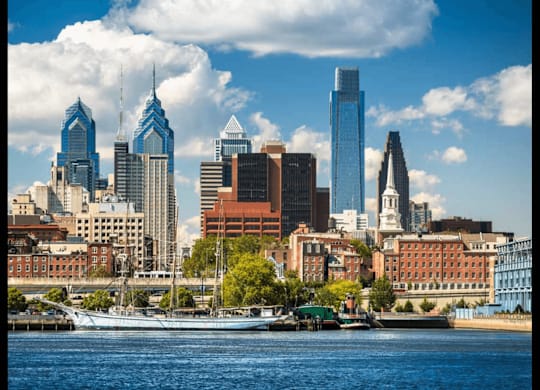 A city skyline with a mix of modern and older buildings, a body of water in the foreground, and a partly cloudy sky.