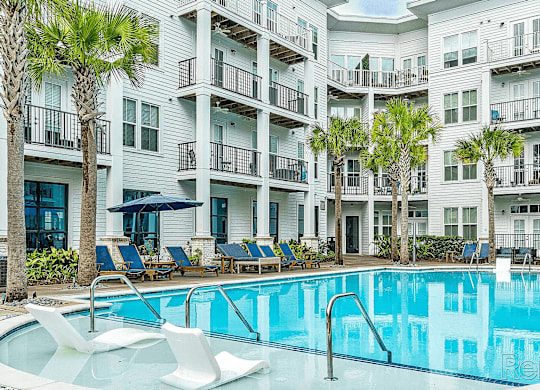 A swimming pool with lounge chairs and palm trees in front of a white apartment building.