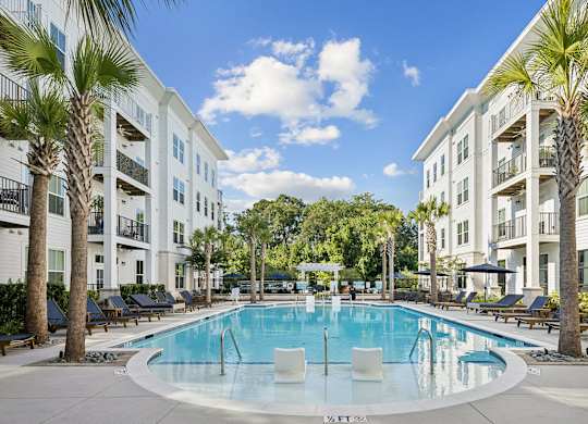 A swimming pool surrounded by palm trees and lounge chairs in front of a white apartment building.