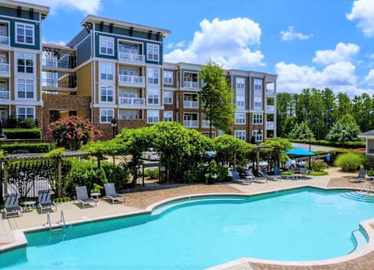 A swimming pool surrounded by lounge chairs and trees in front of apartment buildings.
