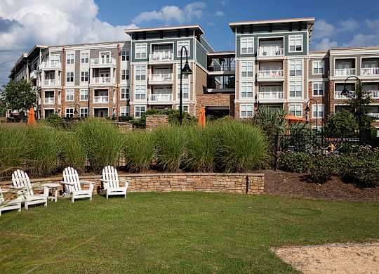 A row of white chairs are set up on a lawn in front of apartment buildings.
