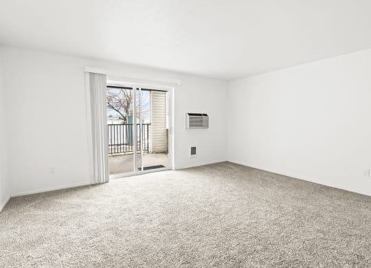 Bright white living room area with taupe carpet, and sliding glass door with vertical blinds at far end of the room..at North Pointe, Post Falls, 83854
