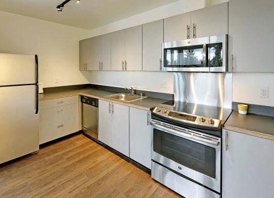 Updated L-shaped kitchen with fridge, dishwasher, sink, oven and microwave from left to right. All appliances are stainless steel and area has wood style flooring.
