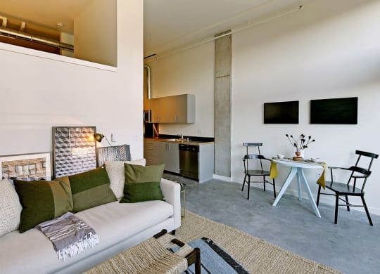 Living room with partial wall behind couch, leading to loft area. Staged with a couch, coffee table, rug and chair. Small dining area at right, with kitchen behind.at 19th & Mercer, Seattle Washington