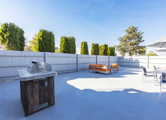 Outdoor lounge with umbrella-covered tables, gas BBQ, and an oversized orange patio couch and coffee table.at The Lakes Apartments, Moses Lake, WA