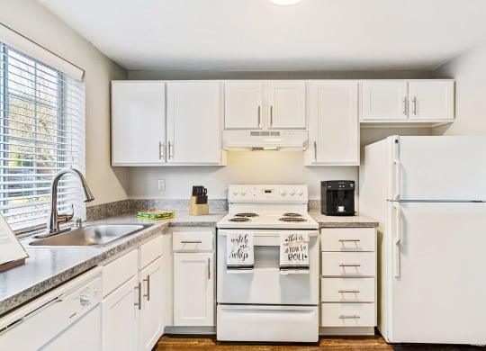L-shaped kitchen with white cabinets, a large window above the sink, and a white dishwasher, oven, stovetop, full-size fridge.at Capitol Crossing, Olympia, WA