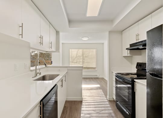 Light-filled galley kitchen with white countertops and cabinets looks into the dining room with sunny windows and white walls. at Pointe East, Fife, WA