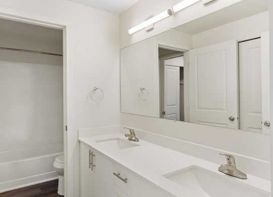 A full bathroom with double vanity sinks, white countertops and cabinets, a large mirror, and a door to the toilet and shower. at Pointe East, Fife, 98424