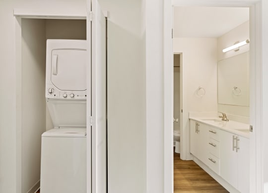 A closet with a white washer and dryer, and the full bathroom to the right with wood flooring. at Pointe East, Fife, WA, 98424