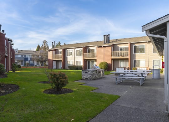 A courtyard with picnic tables, a BBQ, and green grass with apartments in the background. at Pointe East, Fife, WA
