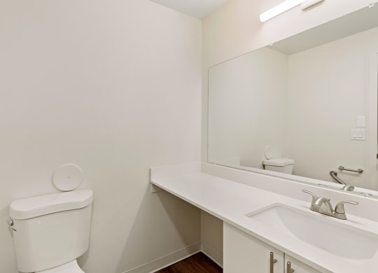 A bathroom with a white vanity sink, under sink cabinets, a large mirror, and a toilet to the left. at Pointe East, Washington