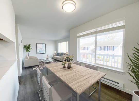 A dining room with white walls and a large sunny window that looks into the living room. Light-filled apartment staged with dining set and living room furniture.at The Trail, Snohomish, WA