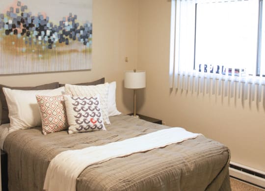Bedroom with queen sized bed and large window with blinds. Staged with bed, side table, lamp and wall art.at The Lakes Apartments, Moses Lake, 98837