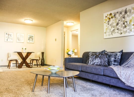 Carpeted living room with hallway that leads to bedrooms. Staged with grey couch, coffee table and art.at The Lakes Apartments, Washington, 98837