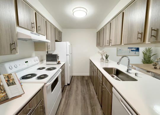 A galley-style kitchen with wood-like flooring, top and bottom cabinet storage, and white appliances. An oven and range, full-size fridge, dishwasher and sink.at The Trail, Snohomish, WA 98290