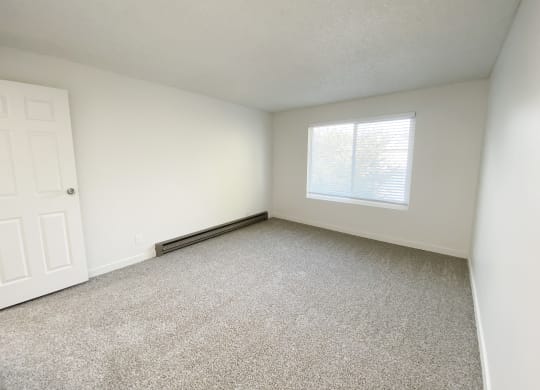 Bedroom with gray carpets, a heater on the left wall, and a large window with blinds on the back wall.at Laurel Park, Twin Falls