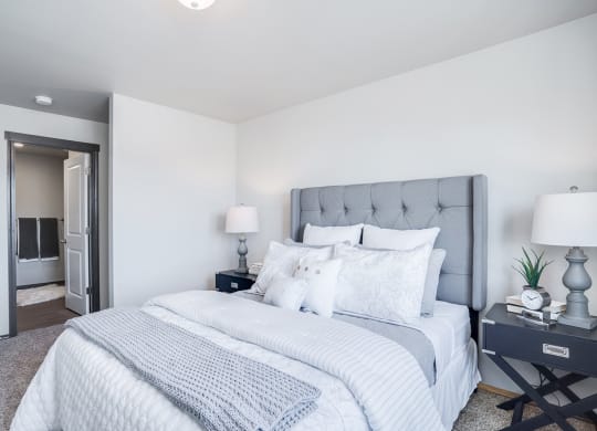 Large carpeted bedroom with full ensuite bathroom on the left. Staged with a queen bed, side tables, and lamps.at North Pointe, Idaho