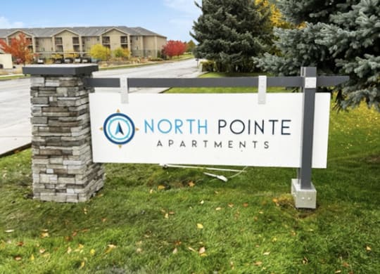 Outside entrance with a blue and white North Pointe sign surrounded by grass, trees, and a road to apartments behind.at North Pointe, Post Falls, ID