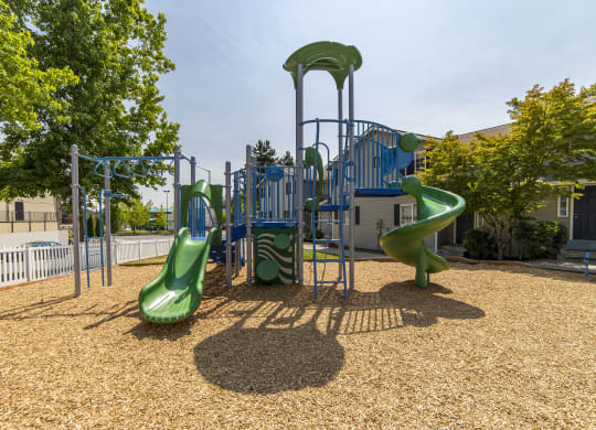 Green and blue playground surfaced with wood chips and enclosed by a white picket fence and green trees at Arterra Apartments, Kent, WA