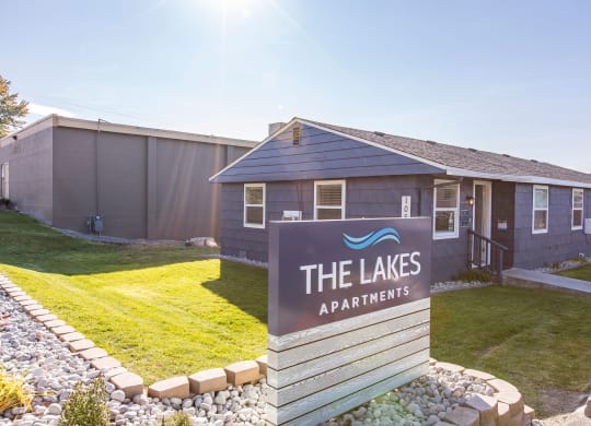 Outside of community with a blue 'The Lakes Apartment' sign surrounded by decorative rocks, green grass, and leasing office behind.at The Lakes Apartments, Moses Lake, WA
