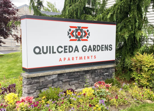 Community entrance with red and gray 'Quilceda Gardens' sign surrounded by flowers, grass, and trees.at Quilceda Gardens, Marysville