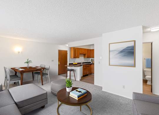 A living room with a grey sofa, a coffee table, a dining table with chairs, a kitchen area with a sink and a fridge, and a painting on the wall.