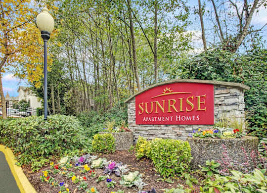 A sign for Sunrise Apartment Homes is surrounded by flowers and greenery.